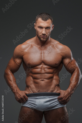 Portrait of a rugged man with a well-groomed model appearance, wearing white briefs, and showcasing his bare muscular torso against a gray background