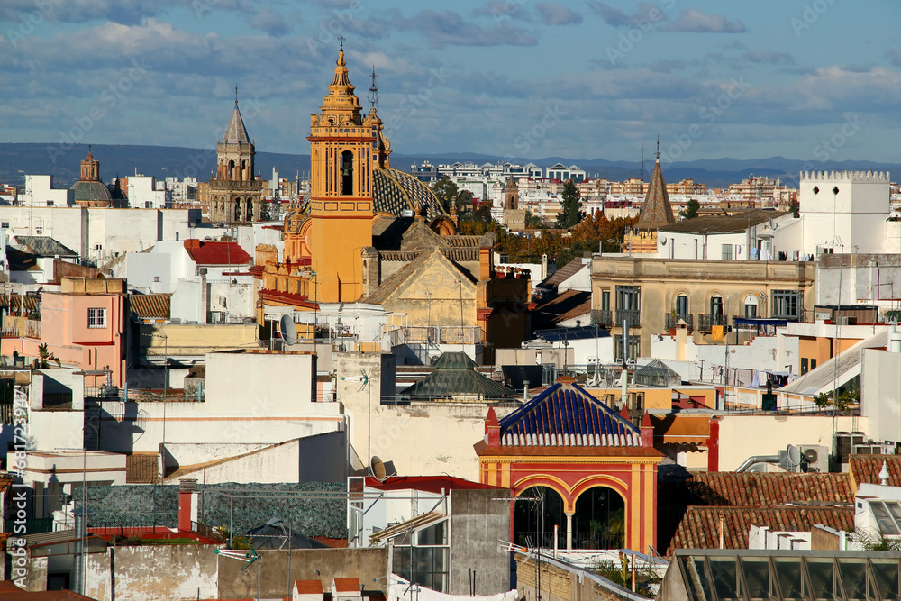View of part of the city of Seville (Andalusia region, southern Spain) with a yellow church in the center of the photo and numerous bell towers around it against a background of a blue sky with clouds
