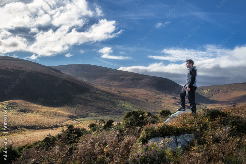 Adult man standing on the rock, illuminated by sunlight, and looking at distant mountains. Hiking in Wicklow Mountains, Ireland
