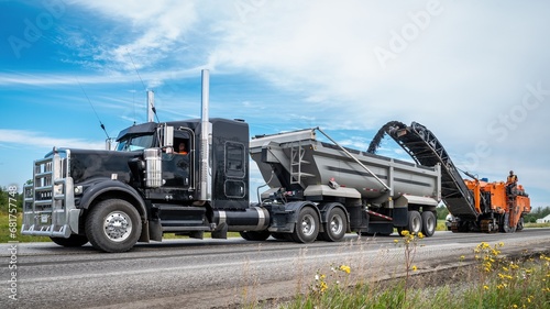 Black semi trailer with a orange planer working on a road construction site, with small flowers in the foreground