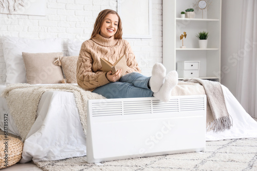 Young woman with book warming legs on radiator in bedroom