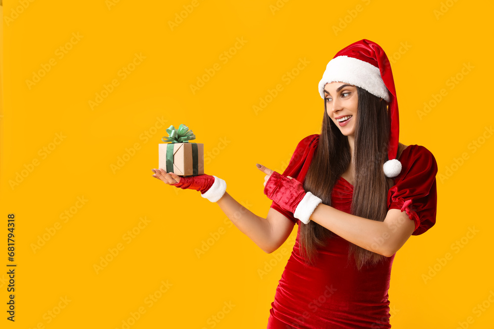 Young woman in Santa hat holding Christmas gift box and pointing at something on yellow background Young woman in Santa hat holding Christmas gift box and pointing at something on yellow background