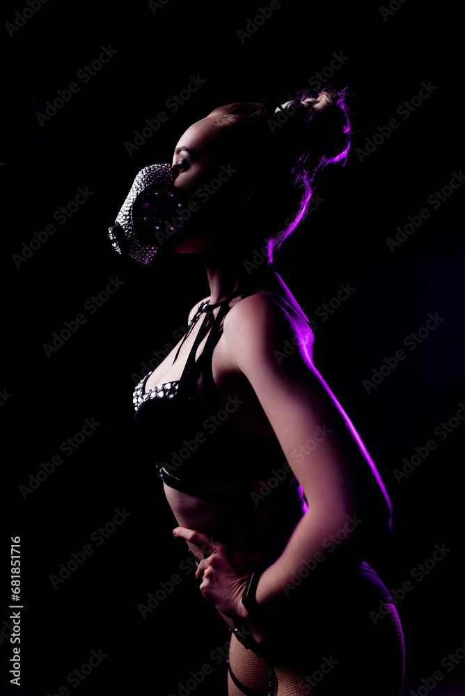 Portrait of a beautiful girl in black underwear and leather harness. On the face is a respirator decorated with rhinestones. Contour purple light. Party concept, night club