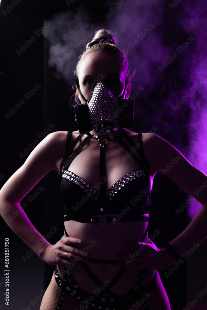 Portrait of a beautiful girl in black lingerie and leather harness. On the face is a respirator decorated with rhinestones. Contour purple light with smoke. Party concept, night club