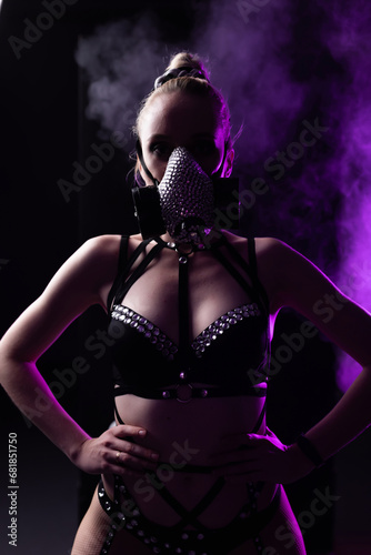 Portrait of a beautiful girl in black lingerie and leather harness. On the face is a respirator decorated with rhinestones. Contour purple light with smoke. Party concept, night club © Granmedia