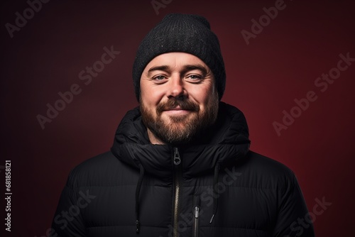 Fat bearded man in a warm hat and jacket on a dark red background.