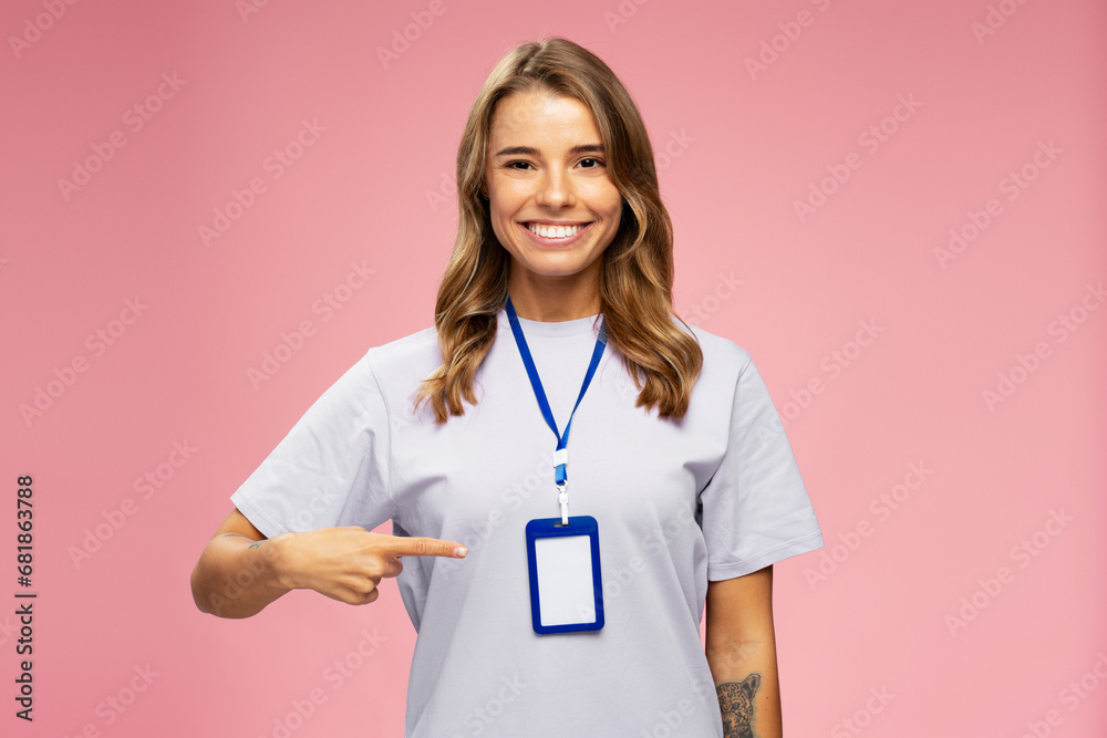 Portrait smiling attractive woman, seller consultant, manager wearing t shirt, pointing on blank badge looking  at camera isolated on pink background. Name card, Mockup  - obrazy, fototapety, plakaty 