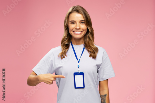 Portrait smiling attractive woman, seller consultant, manager wearing t shirt, pointing on blank badge looking  at camera isolated on pink background. Name card, Mockup  photo