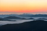 霧に覆われた山脈：オレンジと黄色のグラデーションが広がる空と、地平線上の薄いピンクの線、そして青と灰色の山々が霧やもやに覆われている、日の出または日没の風景