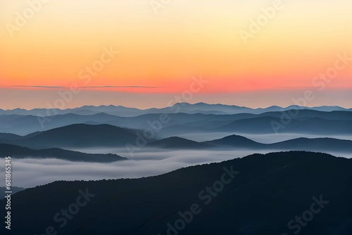 霧に覆われた山脈：オレンジと黄色のグラデーションが広がる空と、地平線上の薄いピンクの線、そして青と灰色の山々が霧やもやに覆われている、日の出または日没の風景 photo