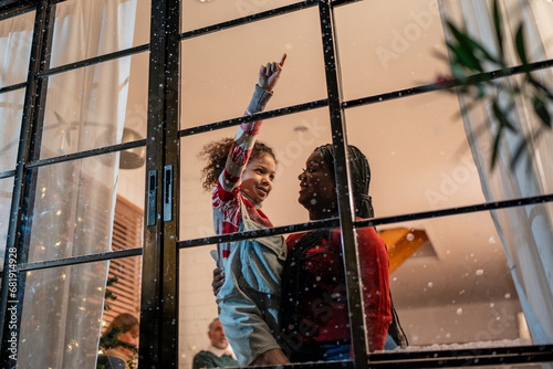 Adorable child looking at the window and first snow flakes with family. 