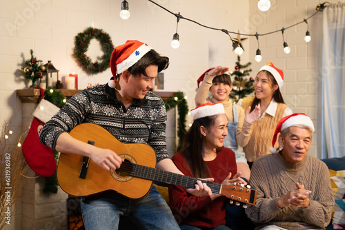 Asian cute family dad playing guitar, mom clap her hand, grandfather sing a song and child girl dance with cousin in Christmas Party night at the living room with festive decoration in hosting home