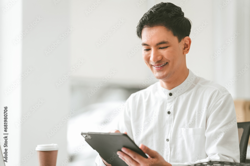 Attractive smiling male asian business person in white shirt working on laptop at desk with coffee cup and documents nearby. Attractive smiling male asian business person in white shirt working on laptop at desk with coffee cup and documents nearby.