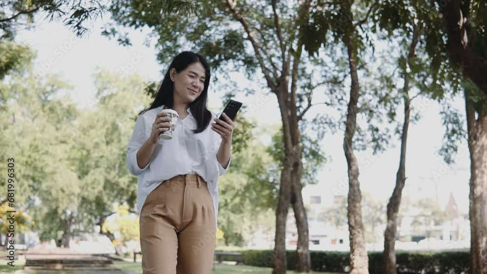 A young Asian woman is walking using a smartphone and drinking coffee in a takeaway cup in the park. Lifestyle and women concept. A young Asian woman is walking using a smartphone and drinking coffee in a takeaway cup in the park. Lifestyle and women concept.