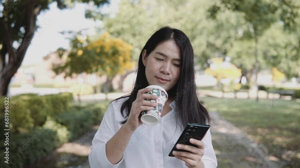 A young Asian woman is walking using a smartphone and drinking coffee in a takeaway cup in the park. Lifestyle and women concept. A young Asian woman is walking using a smartphone and drinking coffee in a takeaway cup in the park. Lifestyle and women concept.