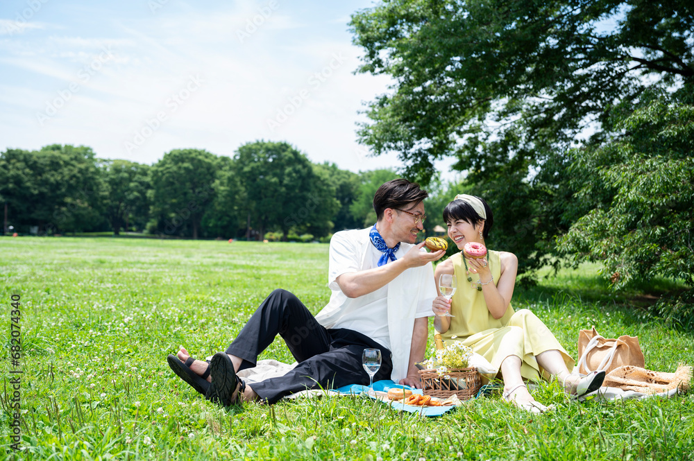 Beautiful Asian couple sitting on picnic mats, smiling happily, eating colourful doughnuts. Beautiful Asian couple sitting on picnic mats, smiling happily, eating colourful doughnuts.