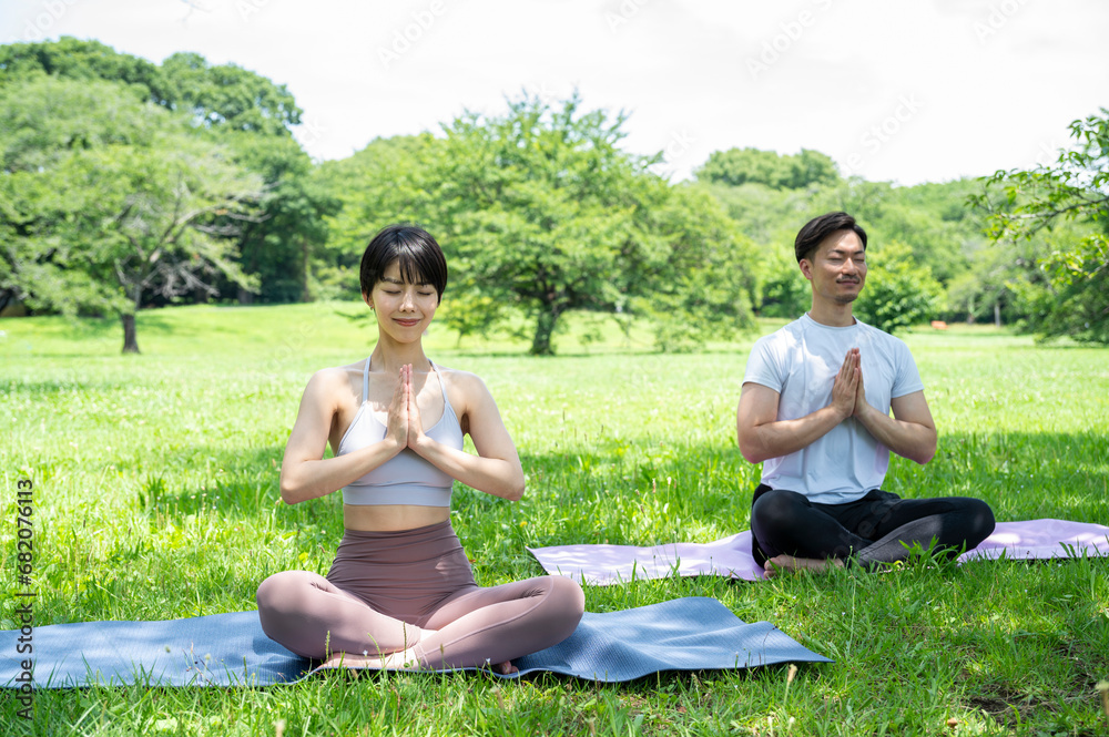 A beautiful Asian couple meditate on a sunny day, pulling out yoga mats in a park with lush green grass. Healthy living, spiritual care, urban living, autonomy, happy lifestyle