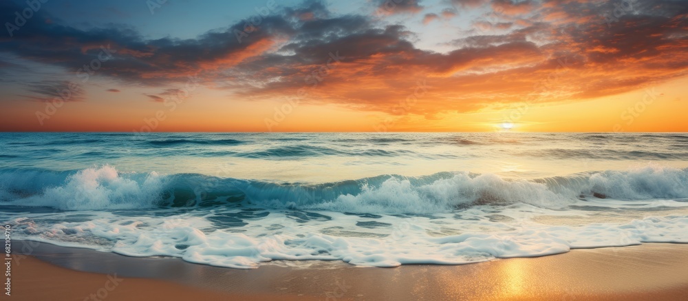As the sun sets behind the horizon, casting a warm orange glow across the beach, the waves crash against the shore, creating a symphony of beauty and tranquility in the summer sky. The light reflects