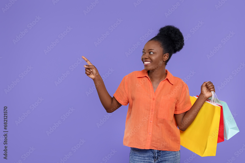 Happy young woman with shopping bags pointing at something on purple background. Space for text Happy young woman with shopping bags pointing at something on purple background. Space for text