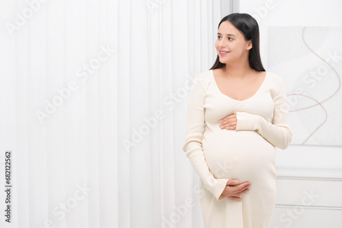 Beautiful pregnant woman in white dress indoors, space for text