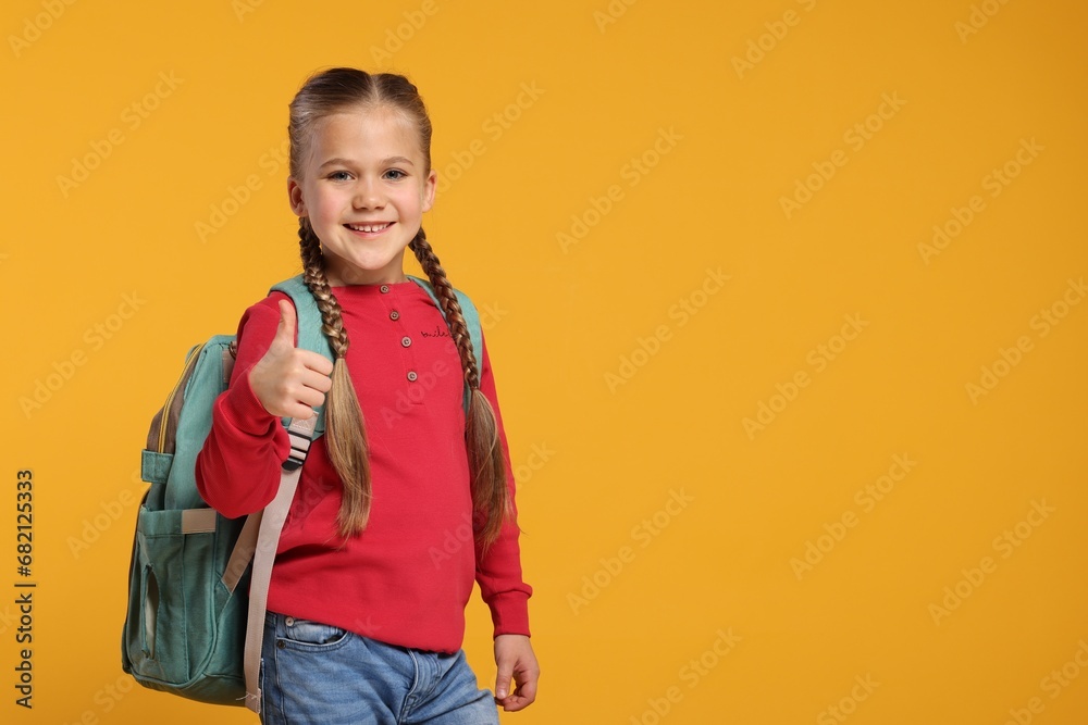 Happy schoolgirl with backpack showing thumb up gesture on orange background, space for text Happy schoolgirl with backpack showing thumb up gesture on orange background, space for text