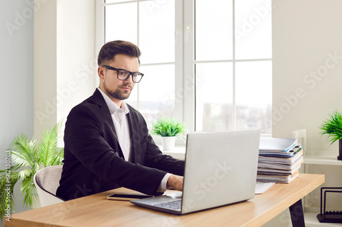 Confident young attractive business man in suit and glasses looking seriously at computer monitor screen working at the desk with a pile of documents on his workplace and typing in the office.