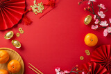 Get glimpse of festive fervor with top view photograph showcasing fans, traditional coins, sycee, Hong Bao, wall hanging, tangerines, sakura on red backdrop, creating space for your text or promotion