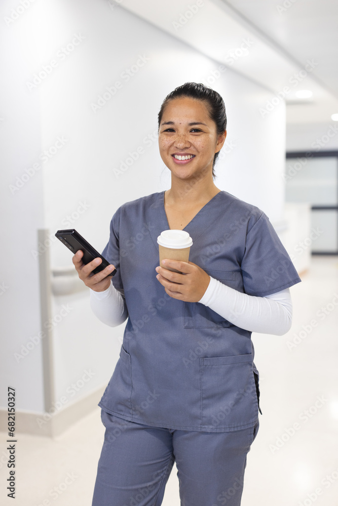 Portrait of happy biracial female doctor using smartphone, holding takeaway coffee in hospital Portrait of happy biracial female doctor using smartphone, holding takeaway coffee in hospital