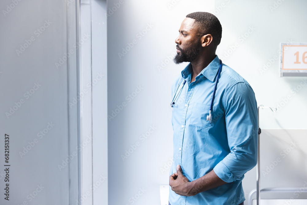 Fototapeta premium Thoughtful african american male doctor looking through window in hospital room, copy space