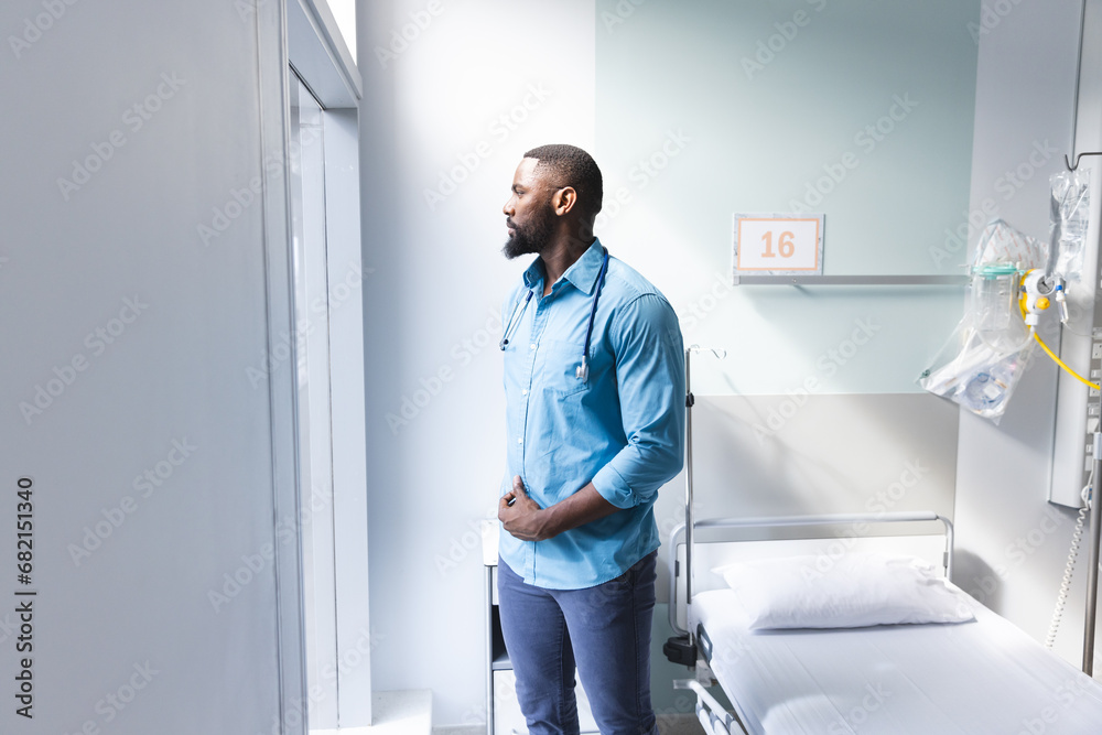 Thoughtful african american male doctor looking through window in hospital room, copy space