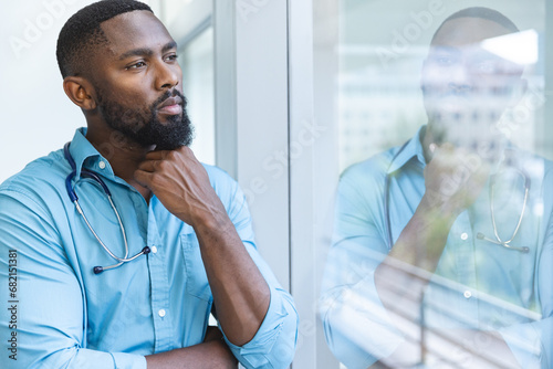 Thoughtful african american male doctor wearing blue shirt looking through window in hospital room