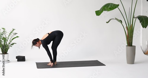 Woman yoga instructor performs Adho mukha shvanasana exercise, downward-facing dog pose with jumping, training in a sports black long-sleeved jumpsuit on a mat in the studio photo