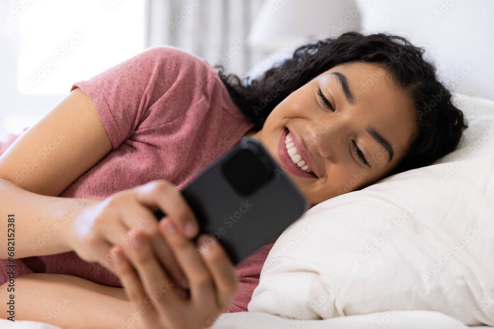 Happy biracial woman using smartphone lying on bed at sunny home Happy biracial woman using smartphone lying on bed at sunny home