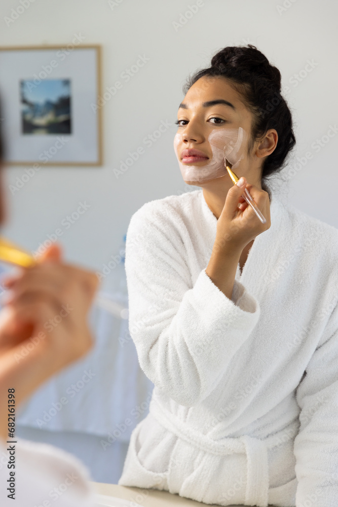 Biracial woman in bathrobe applying face mask using brush in sunny bathroom