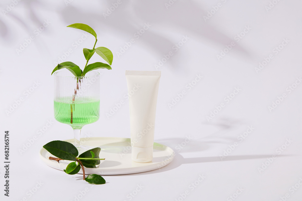 A white cosmetic tube and green tea leaves are displayed on a ceramic plate. The glass contains a transparent green liquid, white background with shadow. Copy space for cosmetics advertising. A white cosmetic tube and green tea leaves are displayed on a ceramic plate. The glass contains a transparent green liquid, white background with shadow. Copy space for cosmetics advertising.