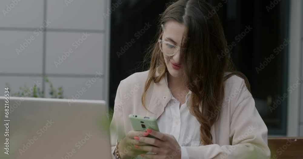 Joyful young woman using smartphone and laughing sitting in street cafe with laptop alone. Modern technology and youth lifestyle concept. Joyful young woman using smartphone and laughing sitting in street cafe with laptop alone. Modern technology and youth lifestyle concept.