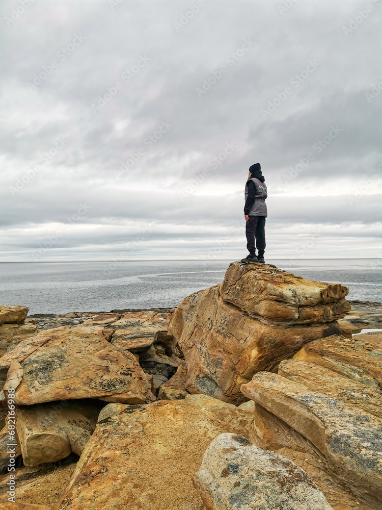 Obraz premium A girl on the shore of red stones next to the rocks of Two brothers on the Fishing Peninsula. The picturesque shore of the harsh Barents Sea. The North of Russia. The Kola Peninsula. The Arctic
