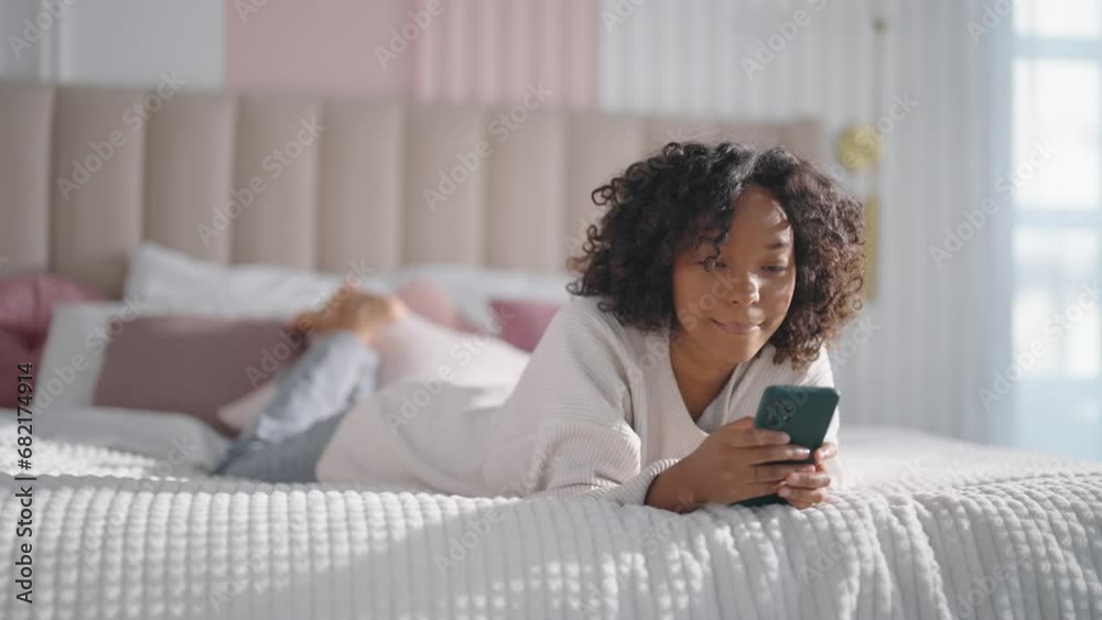 Smiling african american curly woman reading message on smartphone chatting with friend lying in bed at home lazy morning. Positive female enjoying communication in social media looks at phone screen. Smiling african american curly woman reading message on smartphone chatting with friend lying in bed at home lazy morning. Positive female enjoying communication in social media looks at phone screen.