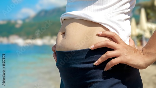 Closeup of young woman with excess fat on belly standing on the sea beach during summer holiday. Concept of health, slimming, loosing weight before summer. photo