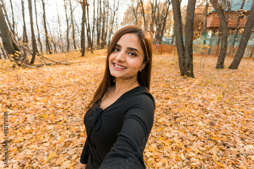 Canvas Print Close-up portrait of diversity young beautiful confident Indian Asian woman in black dress in fall outdoor taking selfie on smartphone for social networks