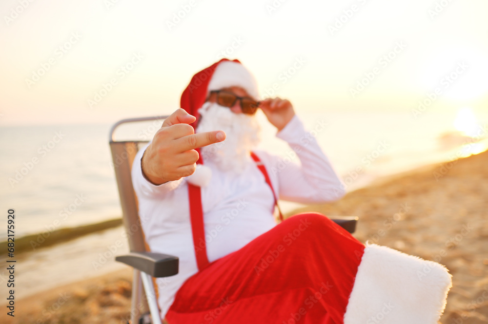 Obraz premium Santa in sunglasses and red suspenders sits in a chair against the background of the sea and the beach and shows the middle finger.