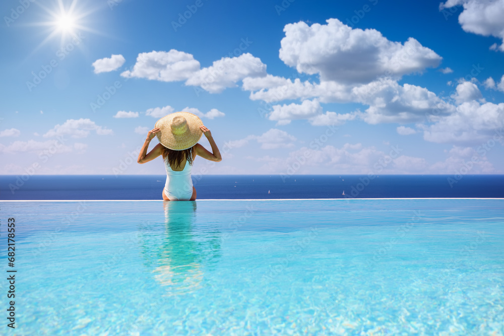 Summer holiday concept with a woman with sunhat sitting at the endge of a big infinity pool - obrazy, fototapety, plakaty 
