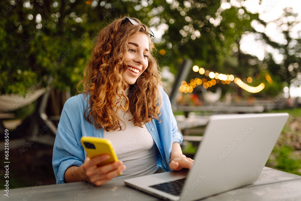 Young curly woman with a phone and laptop sitting at a table against the backdrop of a lake. Female freelancer works at a laptop outdoors. Technology concept. Lifestyle. Young curly woman with a phone and laptop sitting at a table against the backdrop of a lake. Female freelancer works at a laptop outdoors. Technology concept. Lifestyle.