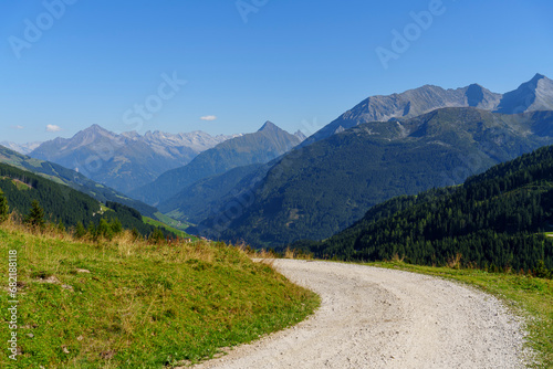 Canvastavla Ein heller Schotterweg windet sich im Vordergrund durch eine grüne Wiese und gibt den Blick frei auf ein weites Gebirgstal mit bewaldeten Hängen und markanten Gipfeln am Horizont