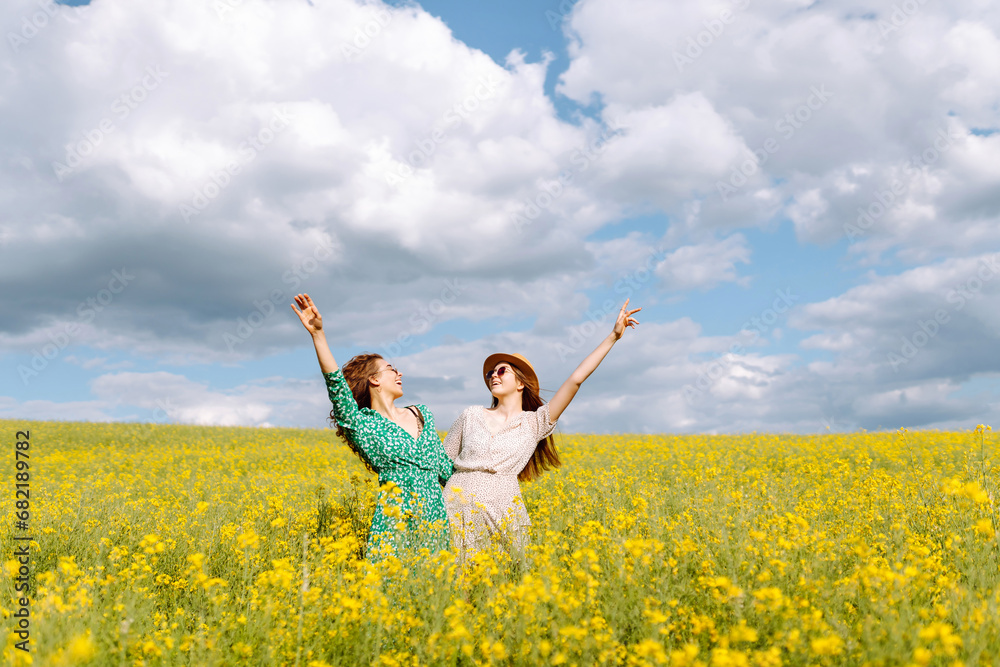 Cheerful girlfriends are walking in a blooming rapeseed field in sunny weather. Two young women enjoying and having fun outdoors. Nature and fun concept. Cheerful girlfriends are walking in a blooming rapeseed field in sunny weather. Two young women enjoying and having fun outdoors. Nature and fun concept.