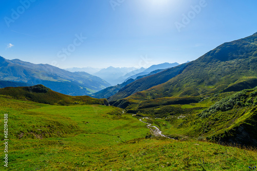 
Ein weiter Blick über saftig grüne Almwiesen in ein tiefes, sonnendurchflutetes Gebirgstal unter einem strahlend blauen Himmel.