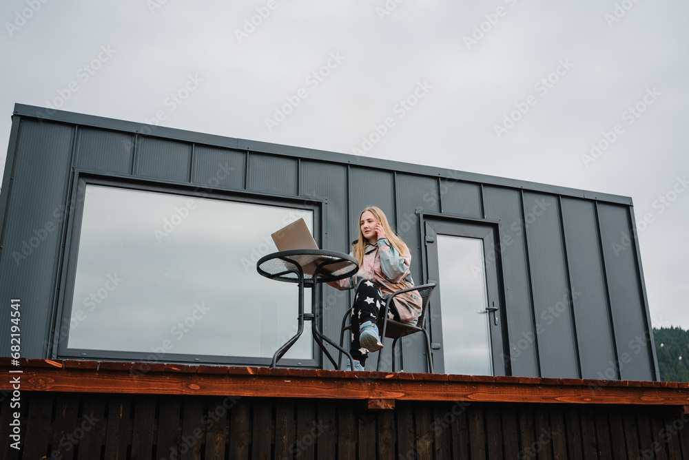 Workplace in country in backyard house in morning. Woman remote work on laptop sitting at table with a great view mountains. Female talking on phone outdoors. Concept freelance lifestyle. Internet 5G.