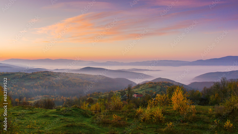 mountainous countryside landscape at dawn. grassy rural fields on the rolling hills in morning light. trees in fall colors. fog in the distant valley. bright sky with clouds above the ridge