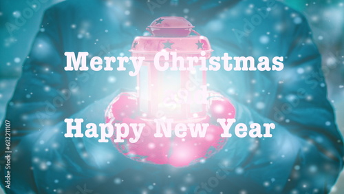 Merry Christmas and Happy New Year words letters design. Woman in jacket and red gloves holding red lantern candlestick with light coming out during snowfall in winter. Text background backdrop.