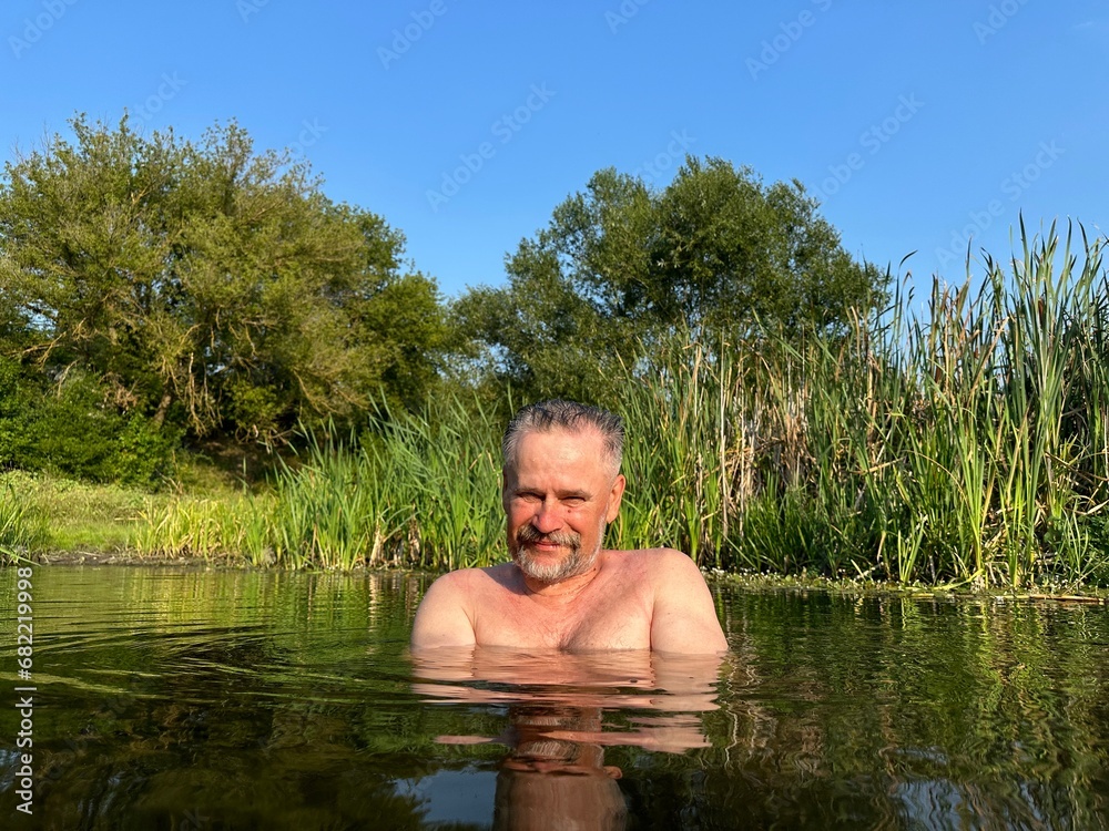 Fototapeta premium Portrait of mature smiling man with gray mustache, standing in dark river water against the backdrop of green reeds and bank with trees, crossing his arms over his chest